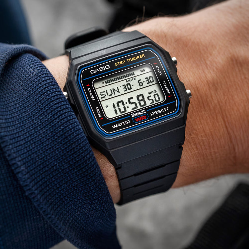 A close-up of a Casio digital wrist watch with a black rectangular case and black strap, worn on a person's wrist. The watch face has blue and orange accents and displays digital time and date information.
