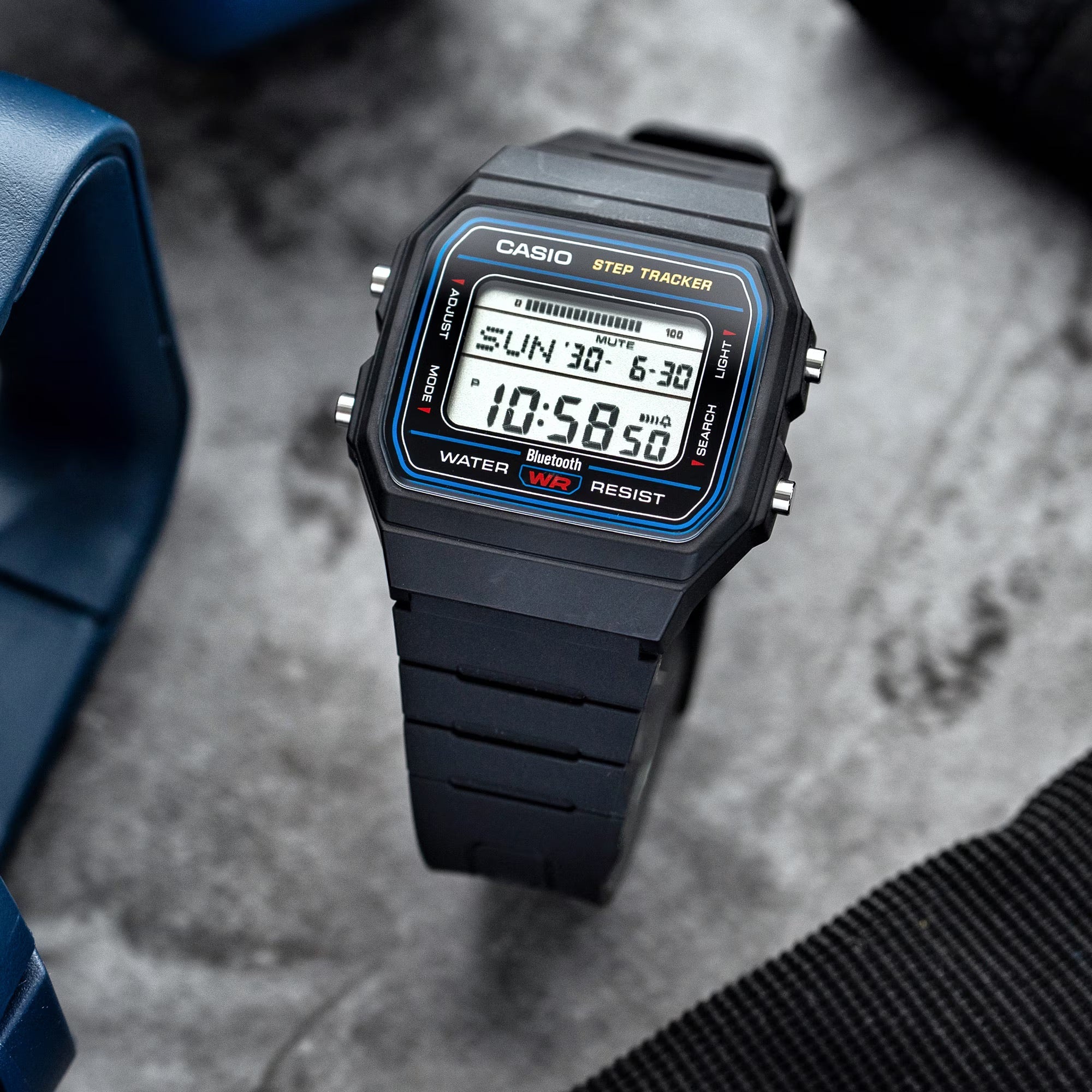 A Casio digital wrist watch with a black rectangular case and black strap, featuring a blue bezel detail and a digital display, photographed on a textured gray surface with partial blue and black props at the edges.