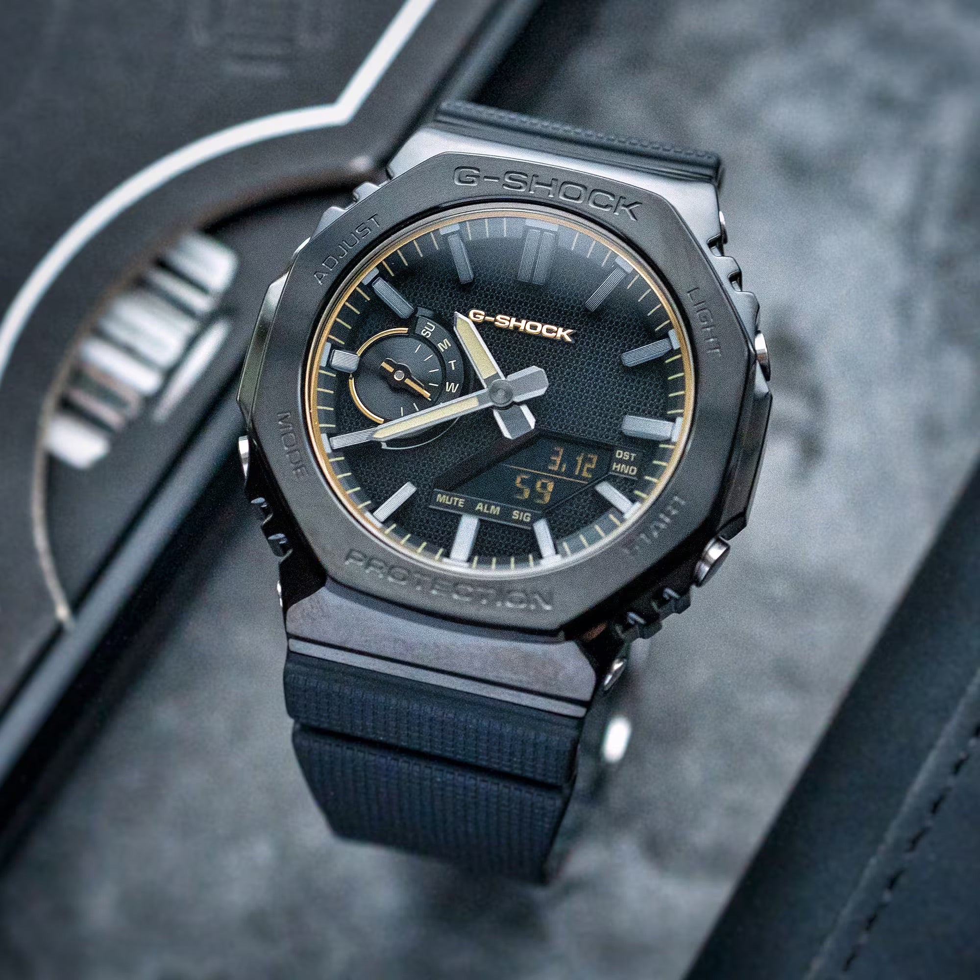 A black G-Shock digital-analog watch with a textured black strap, gold-tone hands and markers, and a dark textured watch face, resting on a dark gray textured surface next to its black presentation box.