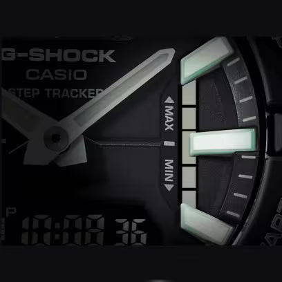 Extreme close-up of a black Casio G-SHOCK watch dial showing luminous green hour markers, silver hands and the step tracker MAX/MIN indicator.