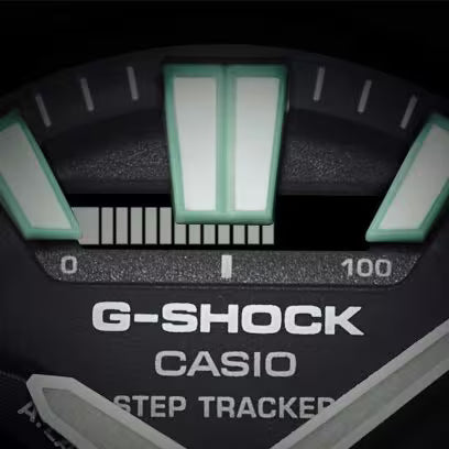 Extreme close-up of a black Casio G-SHOCK watch dial showing green luminous hour markers, silver hands and the step tracker indicator.