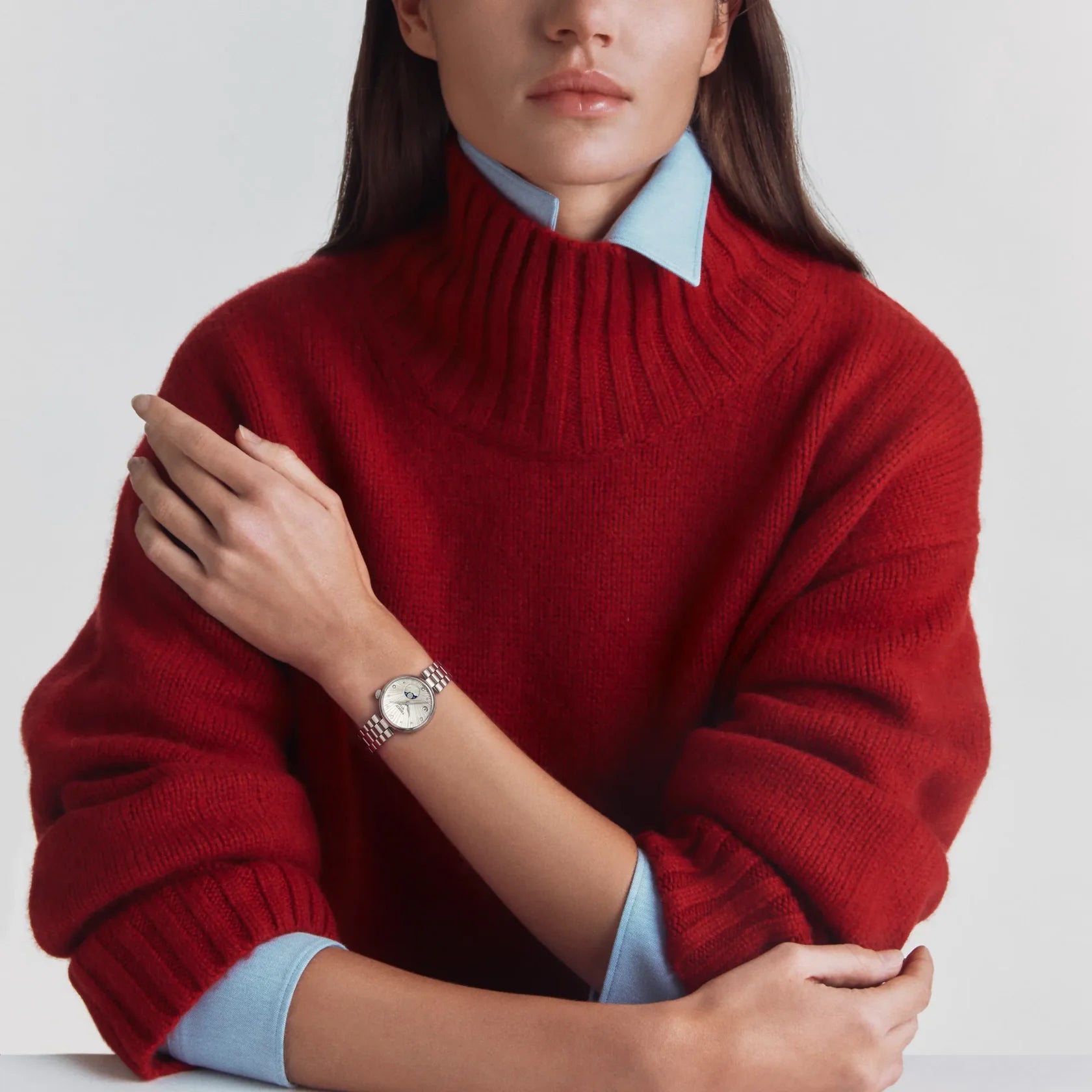 A woman wearing a chunky red ribbed knit turtleneck sweater over a light blue collared shirt, with a silver watch visible on her wrist, set against a clean light gray background.